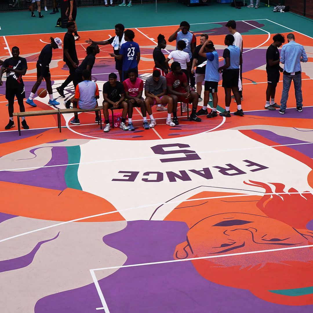 A group of people are sitting and standing on a basketball court, which features an illustrative artwork, creating a visually engaging scene.
