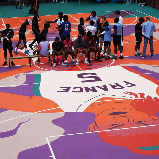 A group of people are sitting and standing on a basketball court, which features an illustrative artwork, creating a visually engaging scene.