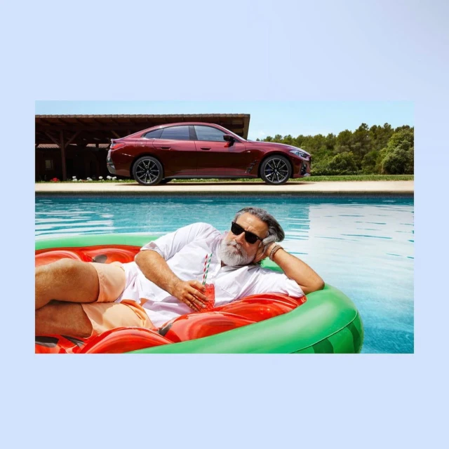 Mature adult man relaxing in a watermelon-shaped float in a pool with a drink, wearing sunglasses; a car and house in the background.