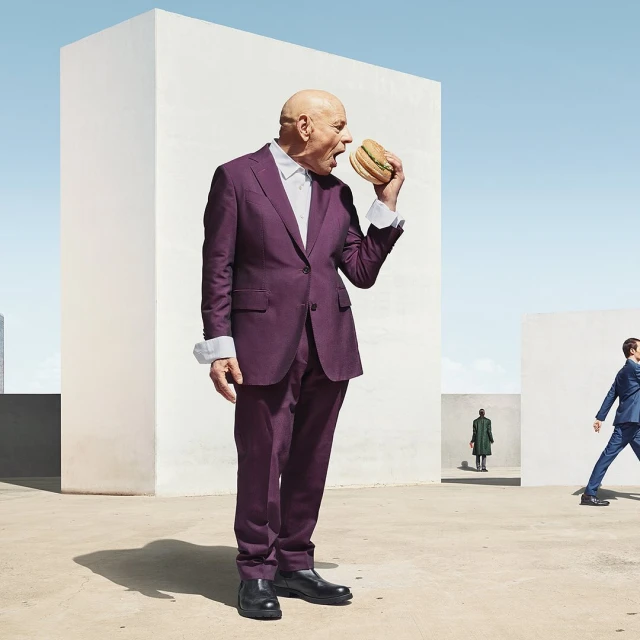 Bald man in purple suit eats burger outdoors, with other figures in background against minimalist architecture on sunny day.