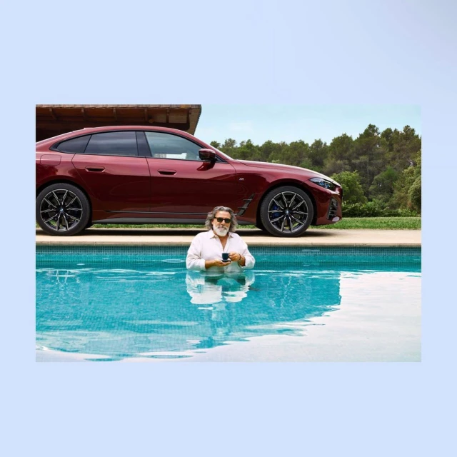 Mature man in a white shirt stands in a pool holding a coffee cup, with a maroon-colored BMW car parked in the background on a sunny day.