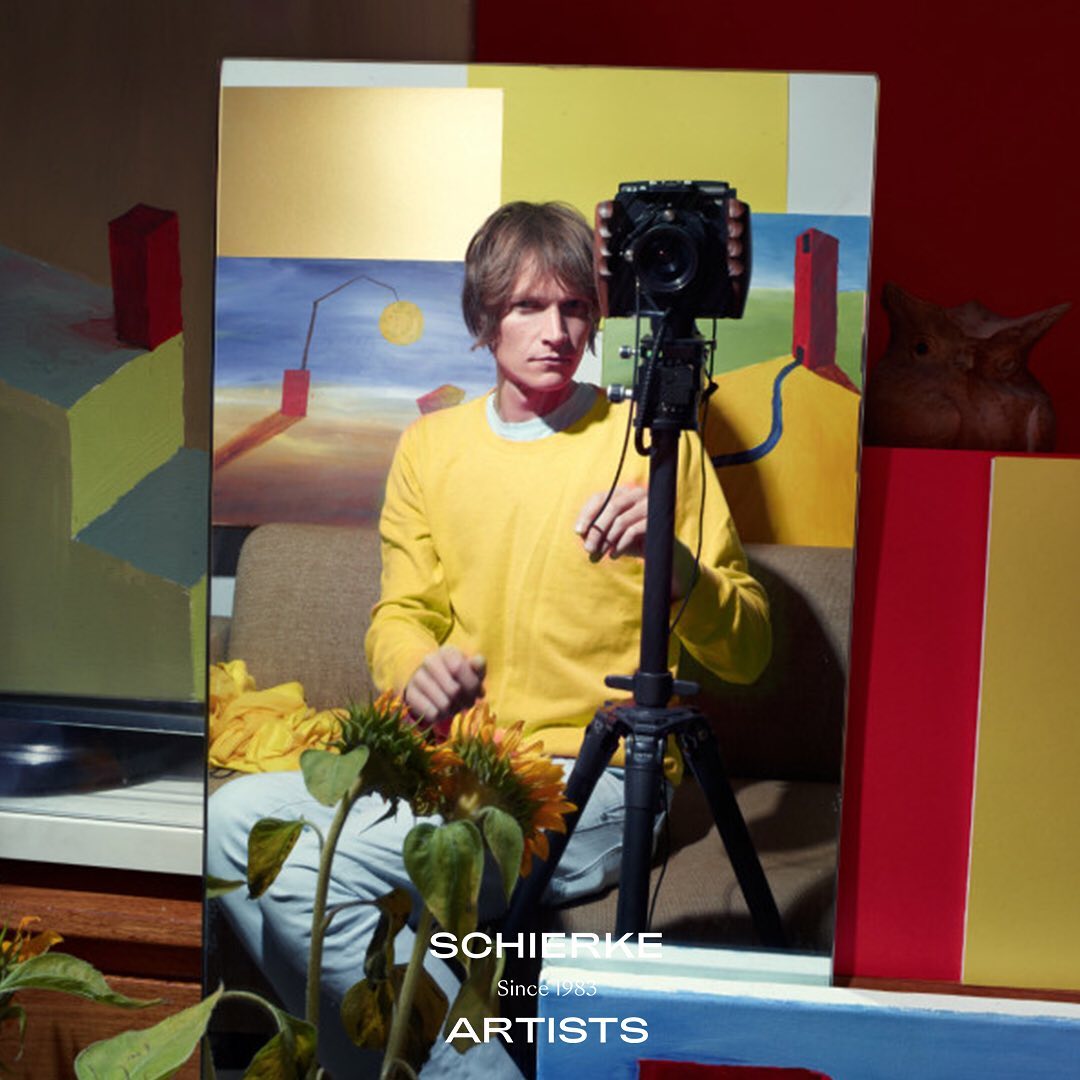 Adult male photographs himself with a DSLR camera mounted on a tripod, sunflowers in foreground, and colorful geometric artwork in background.