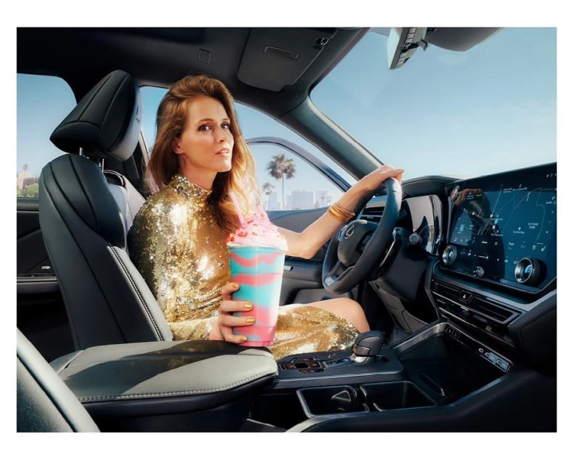 Adult female driving car in urban setting with blue sky and holding frozen drink. Woman in gold dress inside a vehicle.