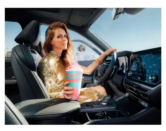 Adult female driving car in urban setting with blue sky and holding frozen drink. Woman in gold dress inside a vehicle.
