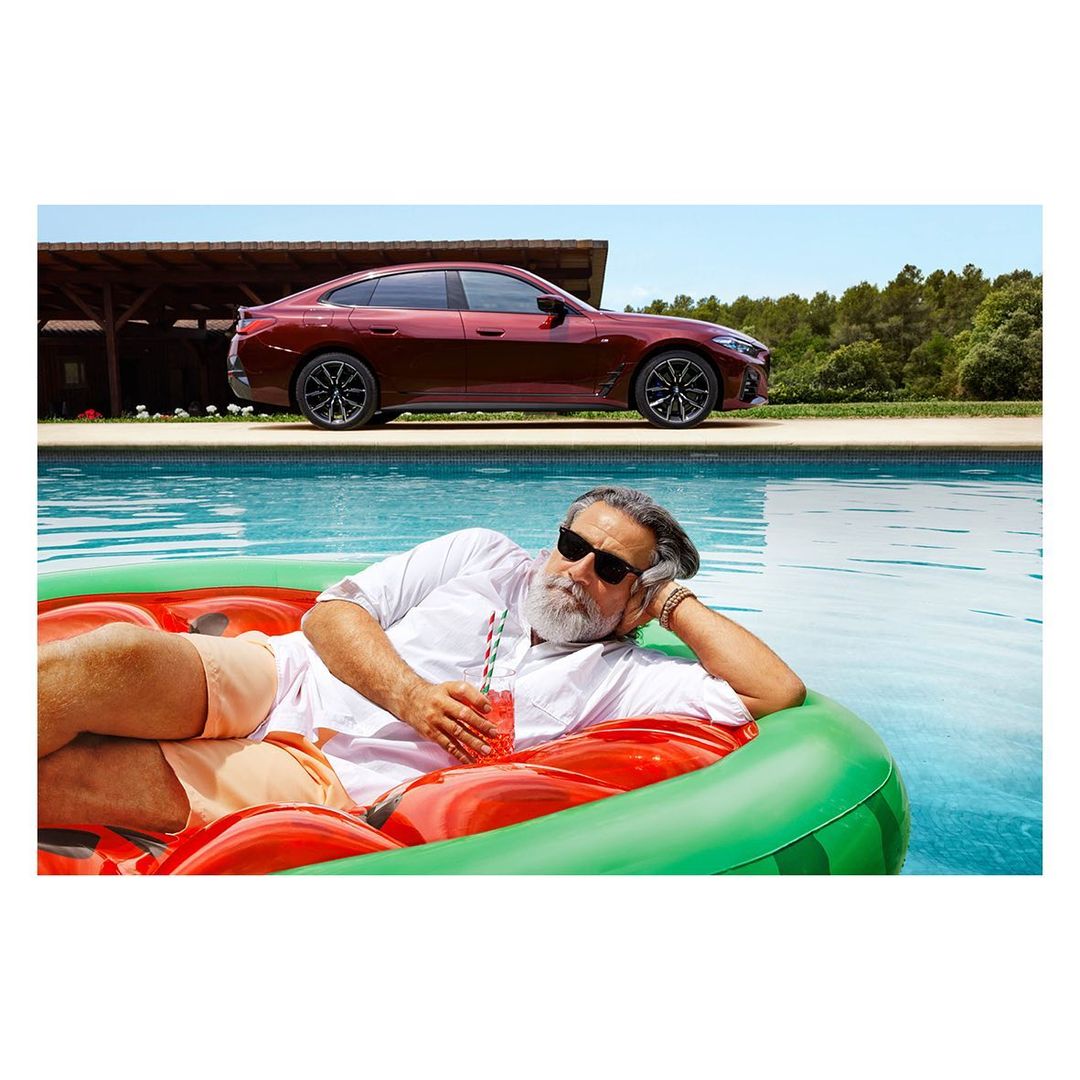 Man in sunglasses relaxes in a watermelon float with a cocktail by a pool, with a dark red car visible in the background.
