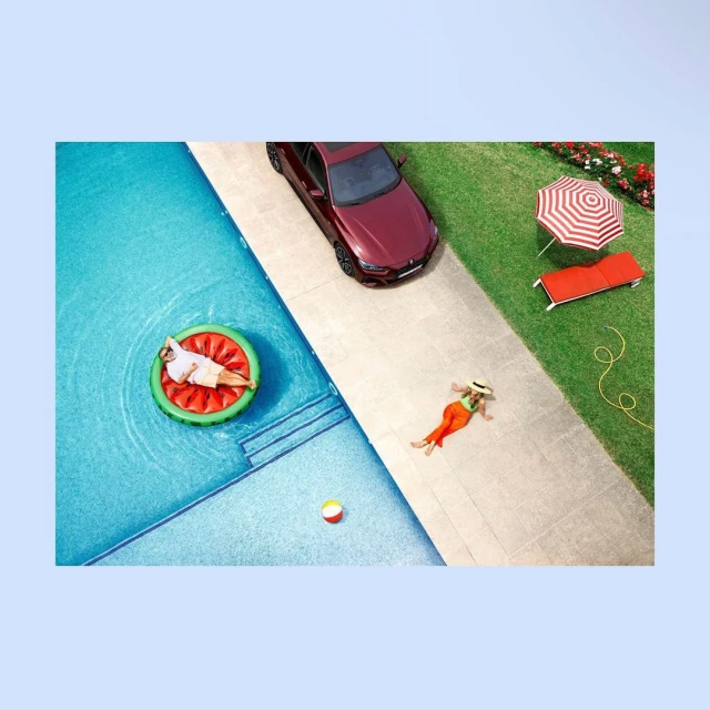 Aerial view of a man on a watermelon float in a pool, a woman sunbathing, a red car, and lawn furniture on a sunny day.