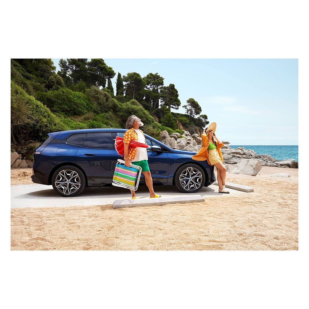A dark-blue BMW parked near an elderly man with luggage, next to a woman leaning on the car at the beach under a clear sky.