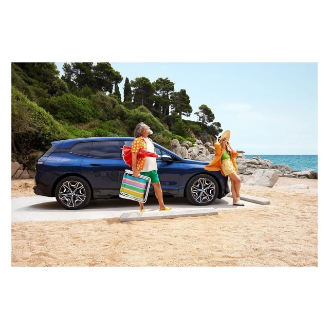A dark-blue BMW parked near an elderly man with luggage, next to a woman leaning on the car at the beach under a clear sky.