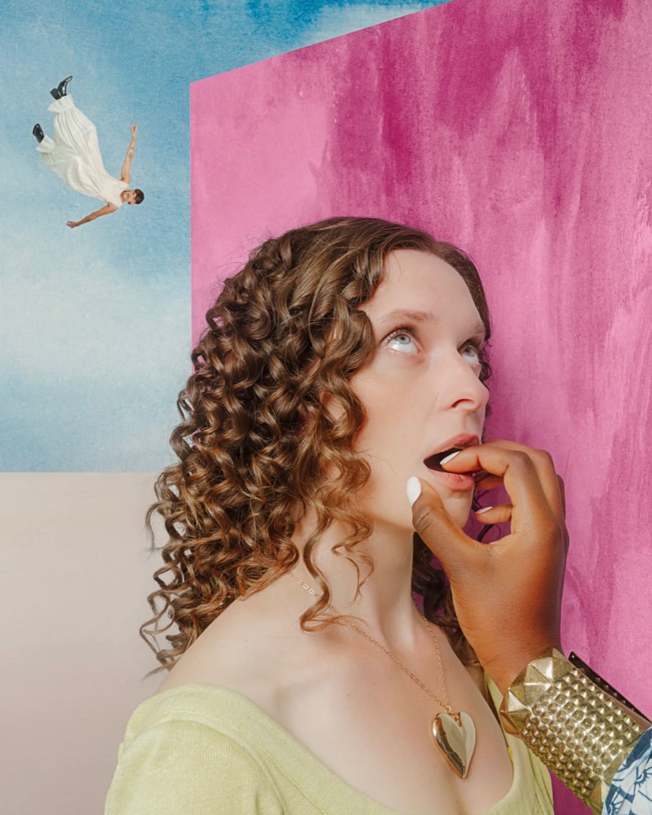 Woman with curly hair is fed a piece of food with an outstretched hand in front of a pink and blue painted backdrop.