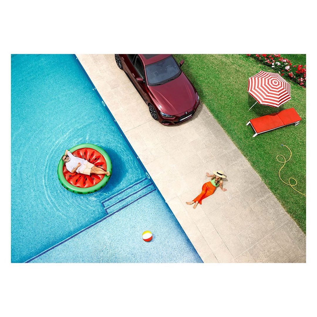 Aerial view captures a man on a watermelon float in a pool, a woman sunbathing, and a parked red car, all under bright daylight.