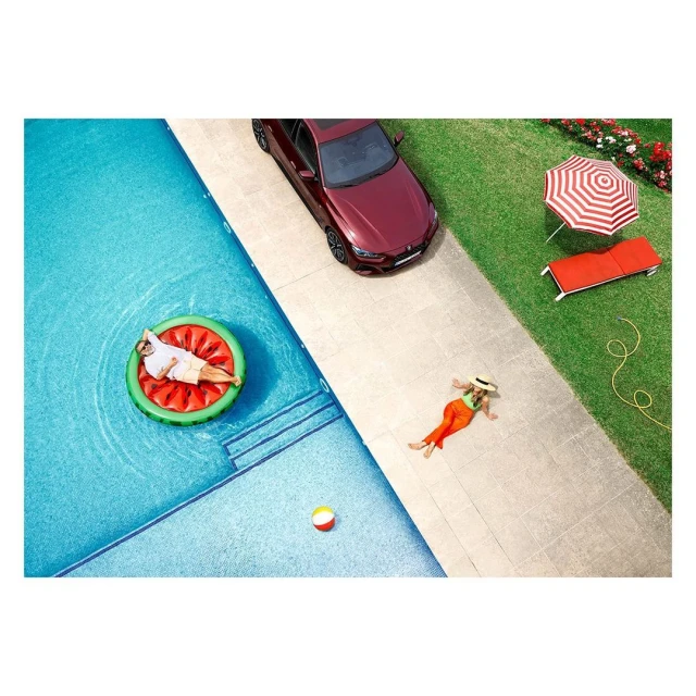 Aerial view captures a man on a watermelon float in a pool, a woman sunbathing, and a parked red car, all under bright daylight.