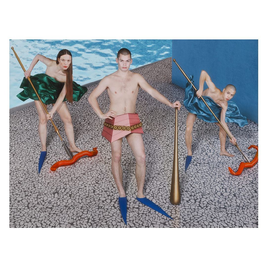 Three figures pose in an artful setting: two women in colorful draped garments wield long poles, and a shirtless man stands centered with a club.