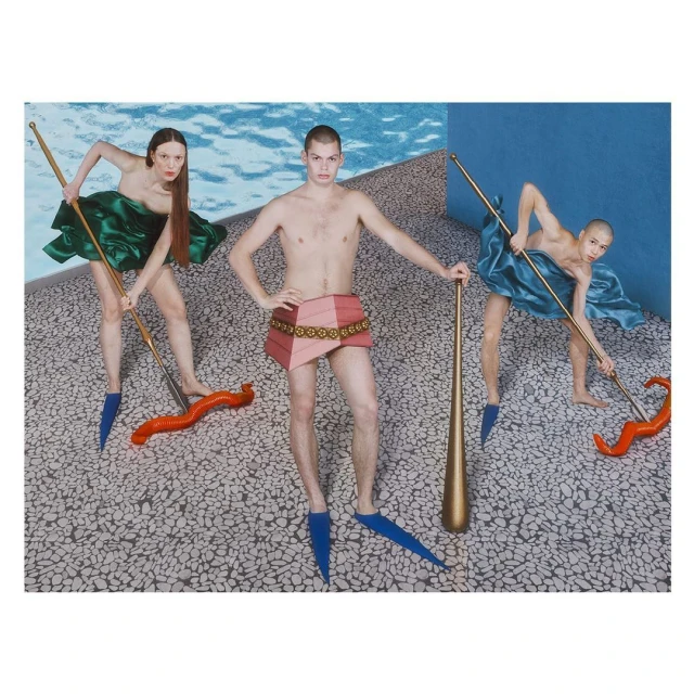 Three figures pose in an artful setting: two women in colorful draped garments wield long poles, and a shirtless man stands centered with a club.