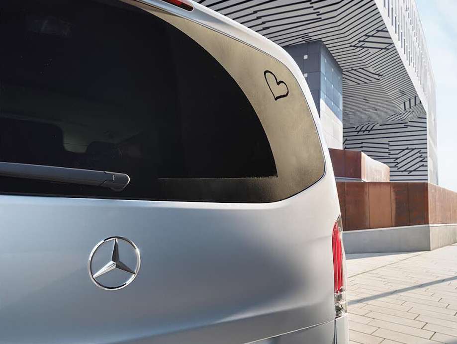 Silver Mercedes-Benz van exterior rear with heart sticker on window, modern architecture in background, neutral daylight.