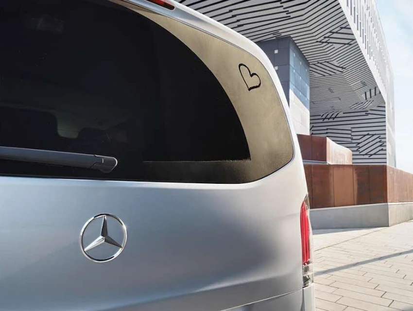 Silver Mercedes-Benz van exterior rear with heart sticker on window, modern architecture in background, neutral daylight.