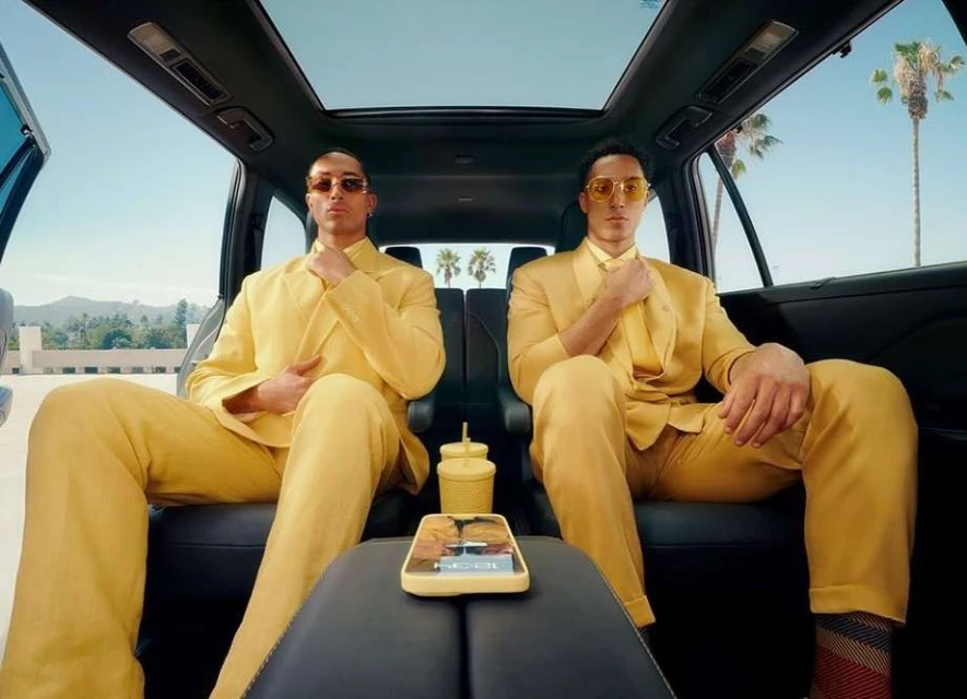 Two men in matching yellow suits sit inside a car in front of palm trees. Drink cups and a cellphone sit on the console.