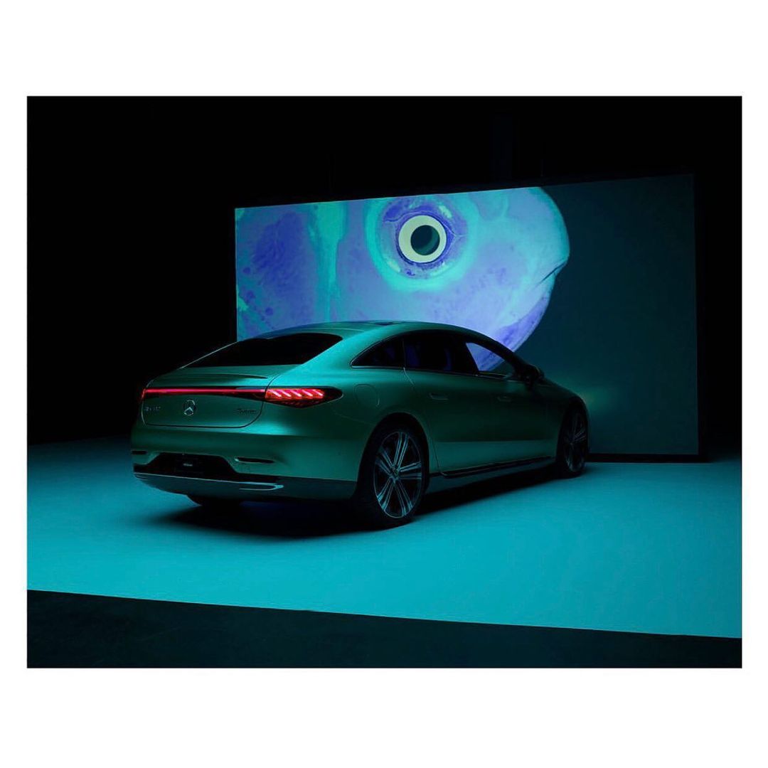 Sleek green Mercedes-Benz EQS coupe, rear view, in a dark studio setting with turquoise floor and abstract eye art projection backdrop.