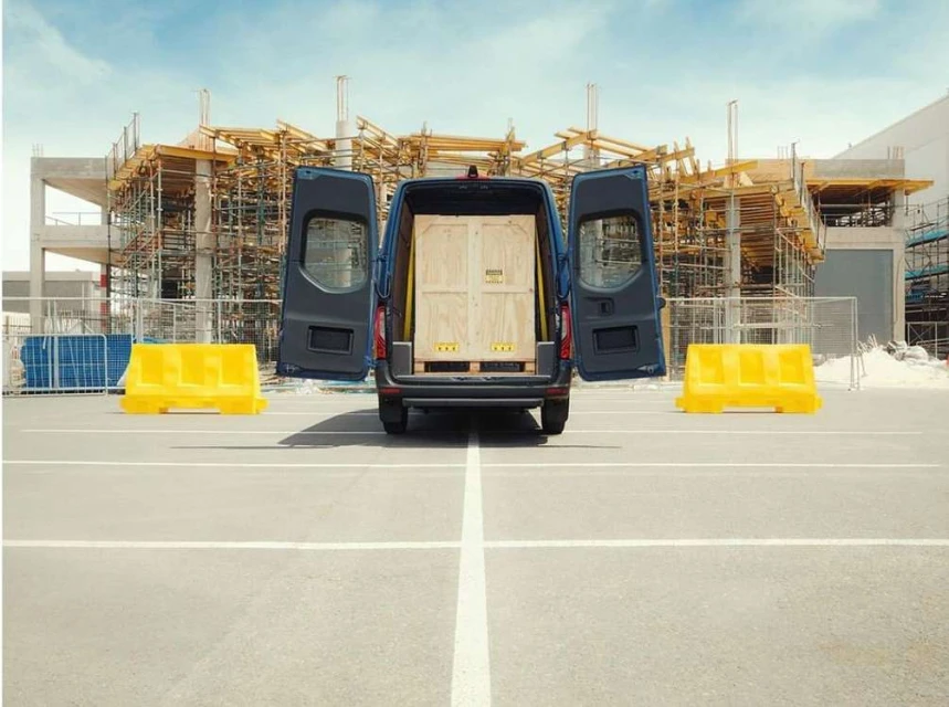 Rear view of a blue commercial van with open doors, revealing a wooden crate inside, parked on a concrete surface with construction site backdrop.
