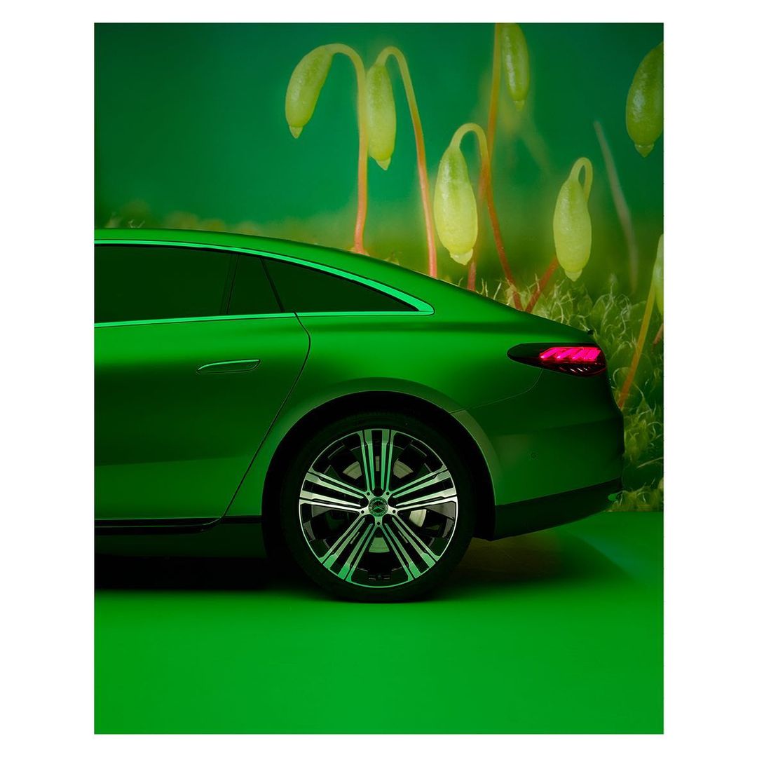 Lime green electric sedan car parked indoors on a green floor, shot at eye level in front of a light green background with out-of-focus plant shapes.