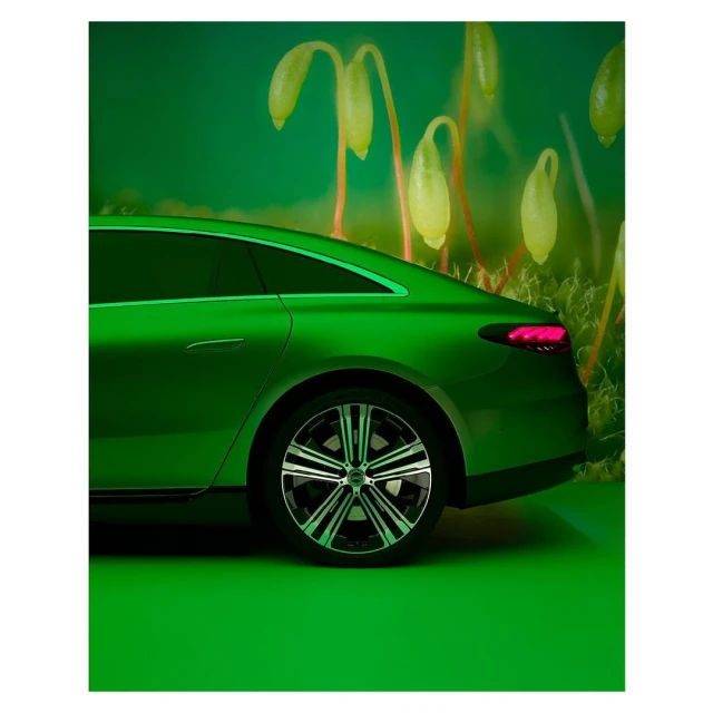 Lime green electric sedan car parked indoors on a green floor, shot at eye level in front of a light green background with out-of-focus plant shapes.