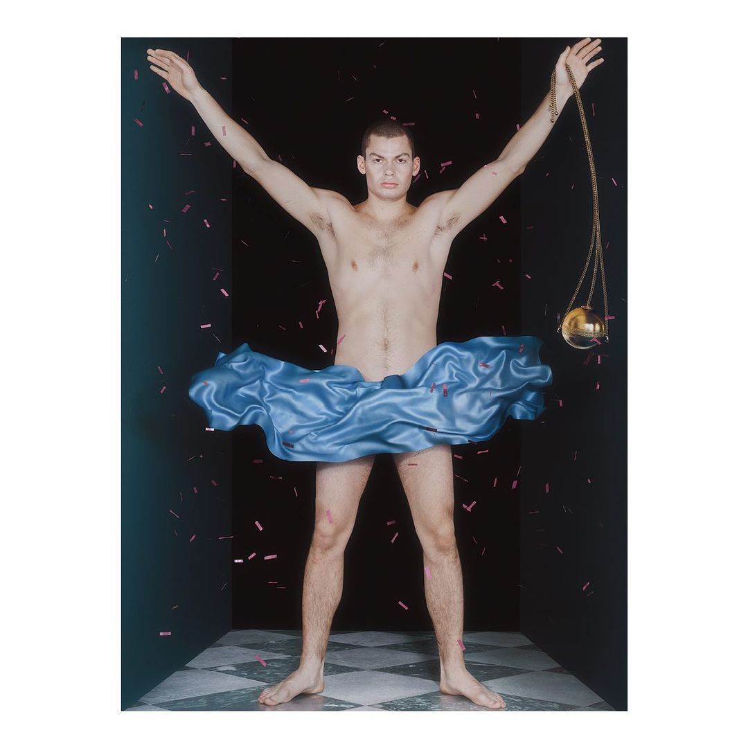 Artistic studio shot of a man in a blue satin skirt with arms raised, confetti falling against a dark backdrop with checkerboard flooring.