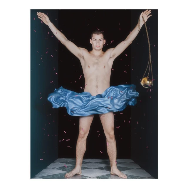 Artistic studio shot of a man in a blue satin skirt with arms raised, confetti falling against a dark backdrop with checkerboard flooring.