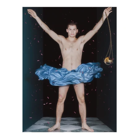Artistic studio shot of a man in a blue satin skirt with arms raised, confetti falling against a dark backdrop with checkerboard flooring.