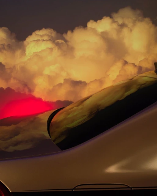 Silver vehicle detail with orange-hued clouds reflected in a window. The vehicle is silhouetted against a red light source.