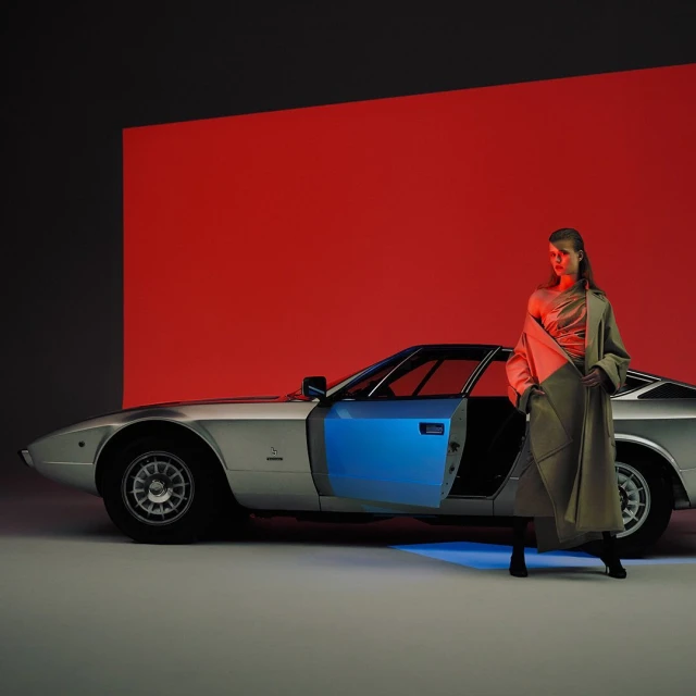 A woman in a tan trench coat stands next to a silver sports car with its door open under red and blue lighting in a studio.