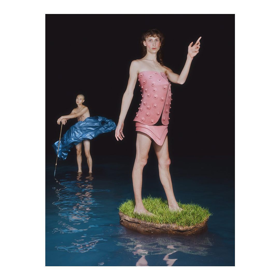 Two figures stand in water, one in a pink outfit on a grassy mound, the other partially submerged with a blue fabric.