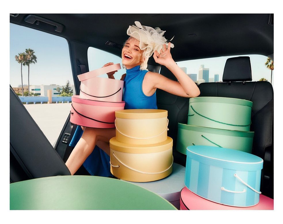 Woman in blue dress and hat surrounded by pastel hat boxes in car with cityscape view, bathed in bright daylight with palm trees.