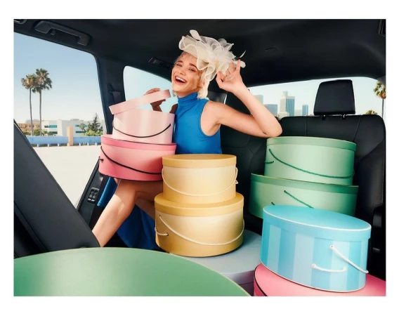 Woman in blue dress and hat surrounded by pastel hat boxes in car with cityscape view, bathed in bright daylight with palm trees.