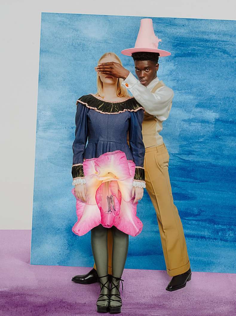 Two fashion models posing in a surreal scene with a denim dress and a large flower, complemented by a light blue backdrop.