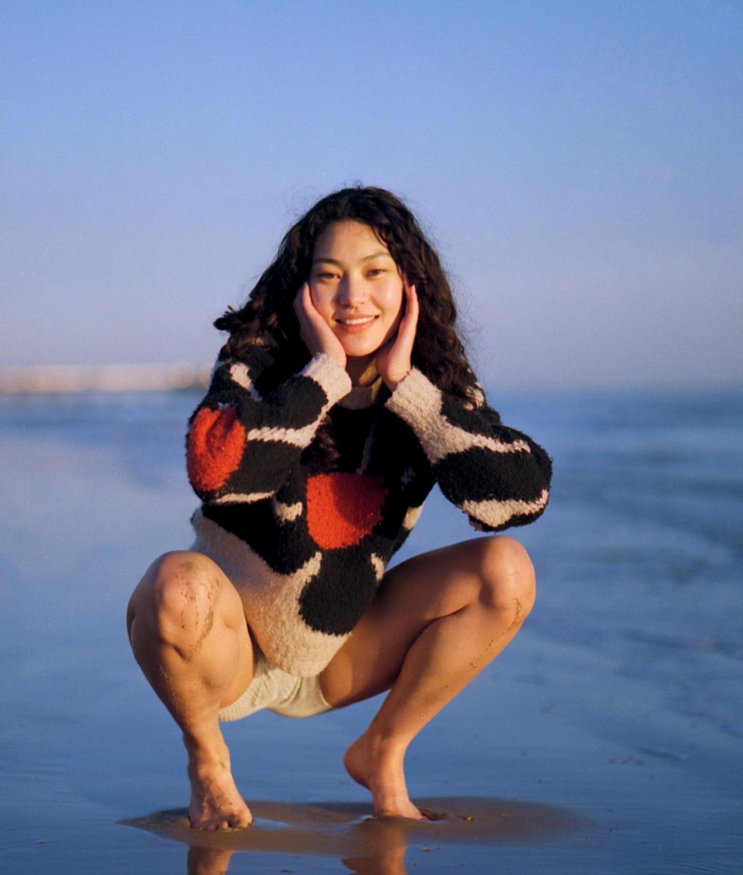 Young woman with dark curly hair squats barefoot at the beach with hands near her face, wearing a black, white, and red sweater.