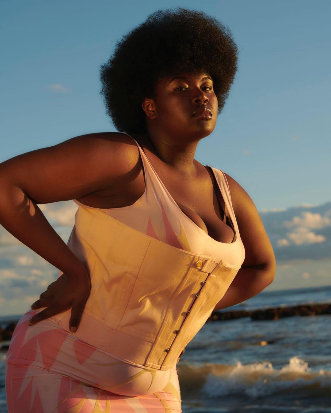Portrait of a plus-size African American woman posing on the beach with a corset and pink patterned skirt in golden hour lighting.