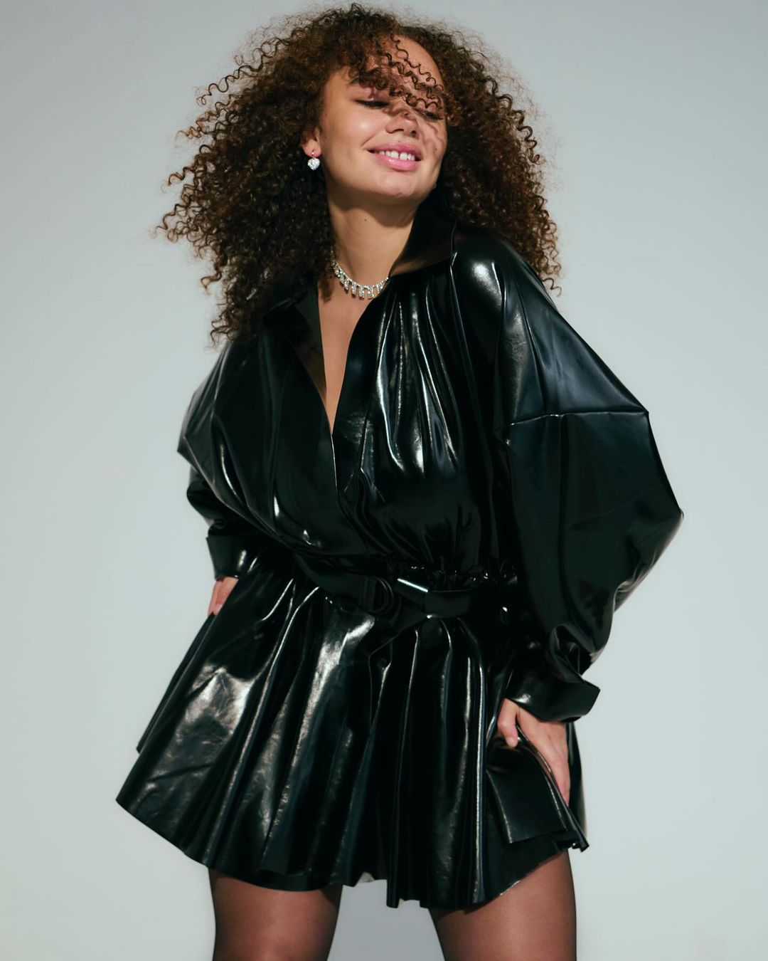 Woman with curly brown hair wearing a glossy black vinyl shirt dress against a pale grey background.