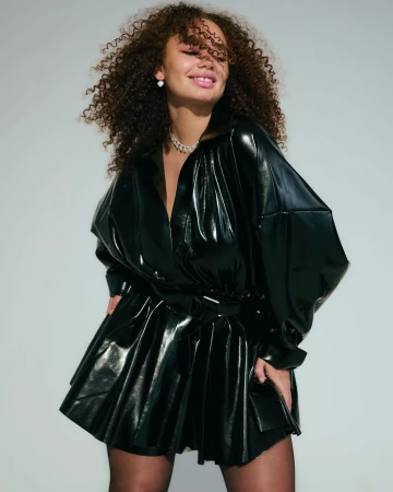 Woman with curly brown hair wearing a glossy black vinyl shirt dress against a pale grey background.