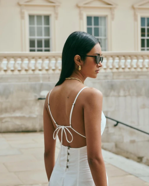 A woman with dark hair in a white dress and sunglasses stands outdoors with a stone wall background, accessorized with gold jewelry.