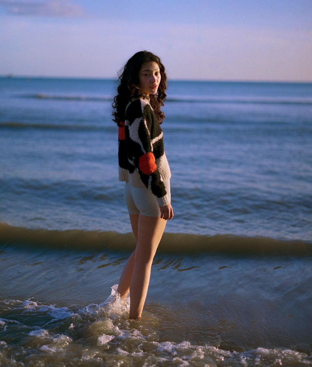 Young woman with curly black hair wearing patterned sweater and light shorts standing in ocean water, looking back over her shoulder.
