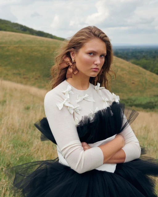 Young woman in a stylish white top with bows and a black tutu poses in a grassy, natural outdoor setting. Portrait orientation.