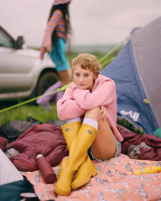 Young woman in pink sweatshirt and yellow boots sits near a tent at an outdoor music festival; a friend is walking nearby.