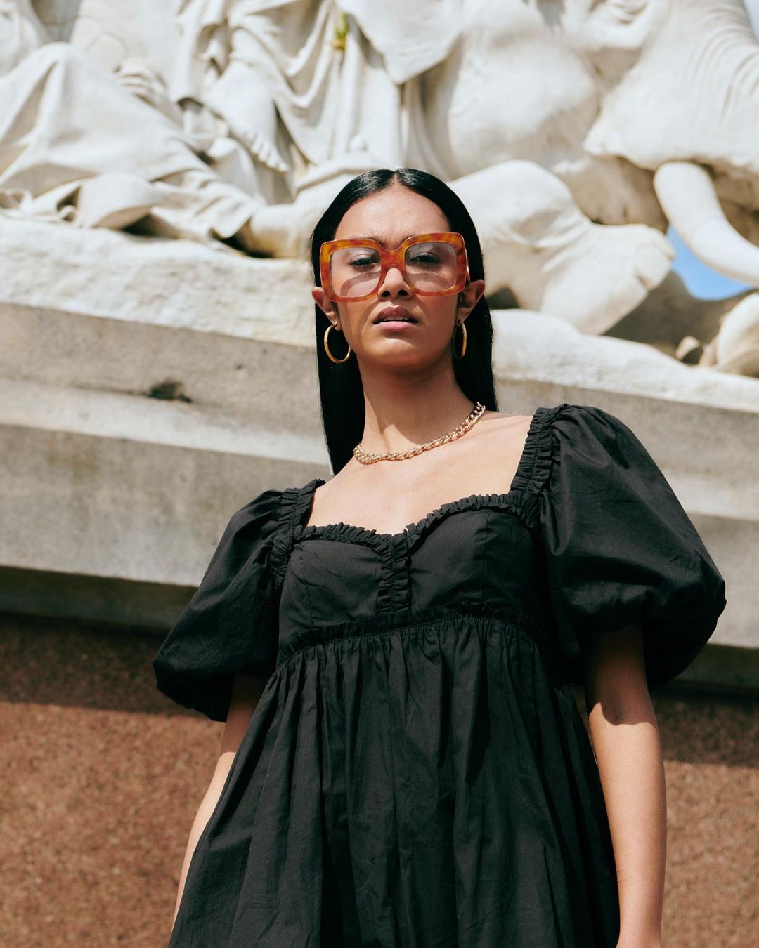 Woman in black dress and large glasses poses in front of an ornate marble sculpture on a sunny day, fashion portrait. 