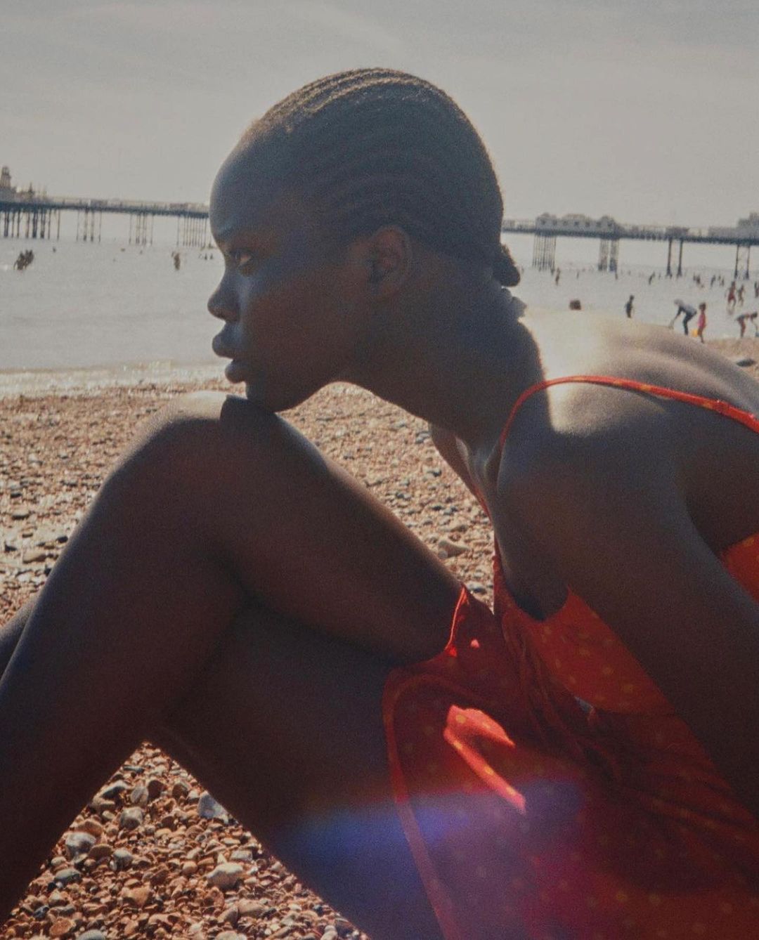 Young woman in red dress sits on a pebble beach near the ocean with a pier visible in the background on a sunny day, profile view.