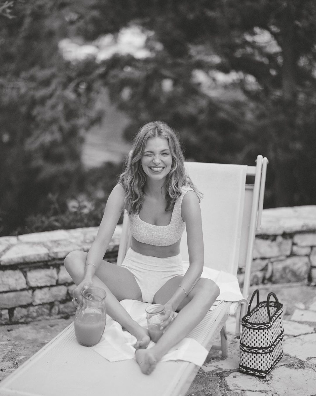 Smiling woman in swimwear sits cross-legged on a lounge chair, holding a drink with pitcher, enjoying a sunny outdoor leisure moment.