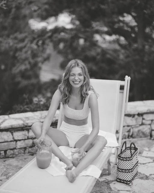 Smiling woman in swimwear sits cross-legged on a lounge chair, holding a drink with pitcher, enjoying a sunny outdoor leisure moment.