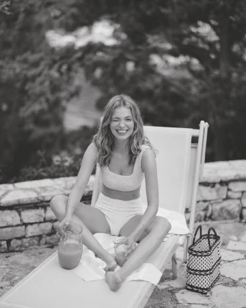 Smiling woman in swimwear sits cross-legged on a lounge chair, holding a drink with pitcher, enjoying a sunny outdoor leisure moment.