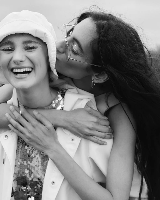 Two women embrace in a black-and-white close-up. One woman kisses the other's cheek as she smiles, wearing a hat and sequined outfit.