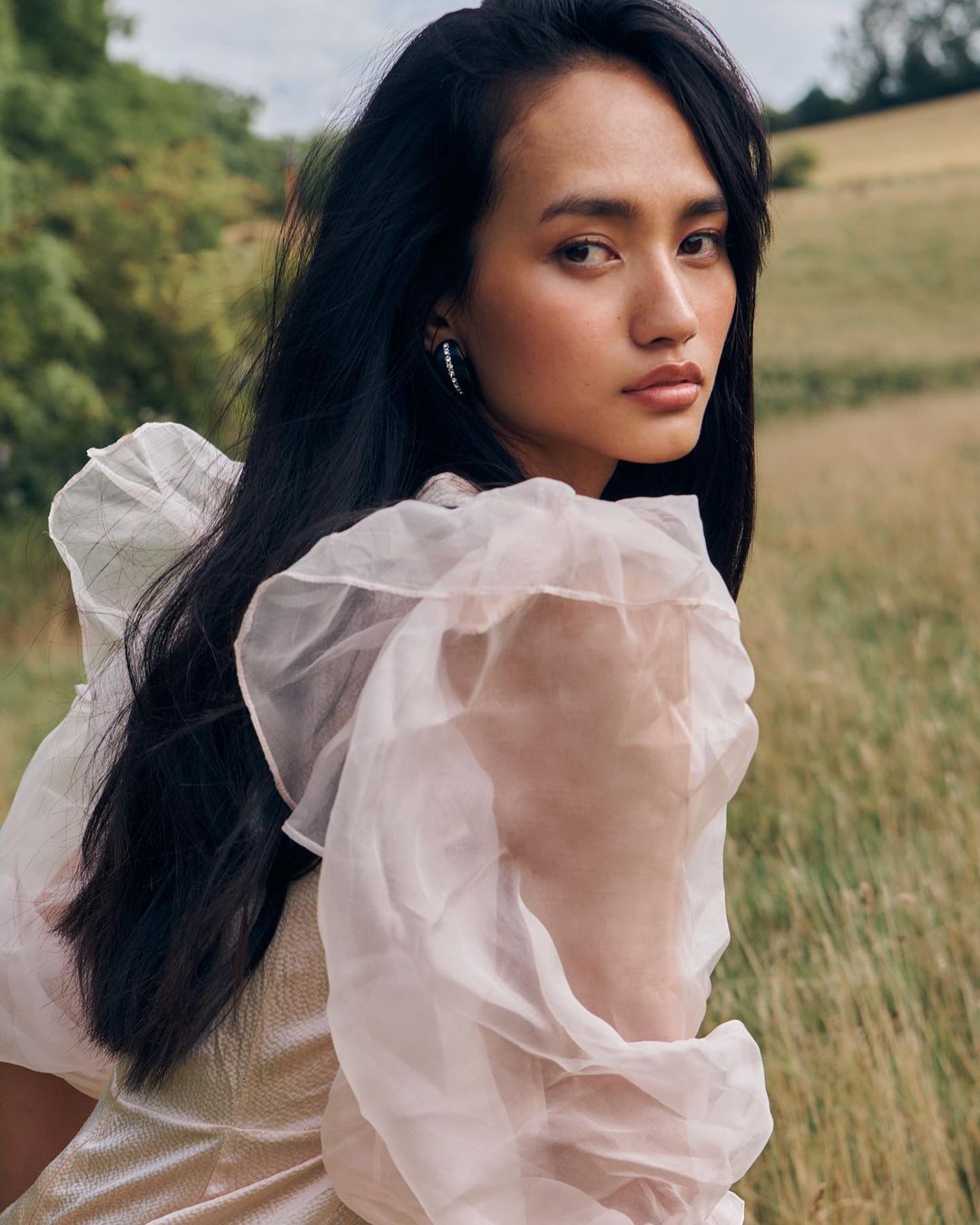 Close-up of an adult woman with long dark hair, wearing a dress with ruffled sleeves, posing outdoors in a field with trees in the background.