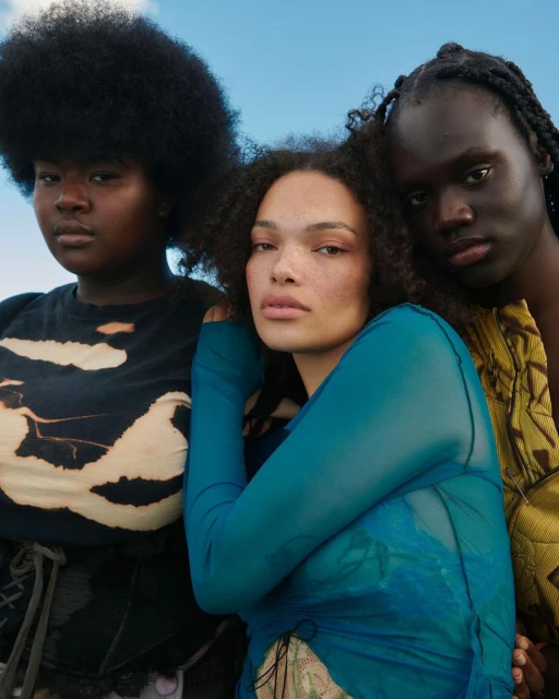 Three diverse adult models pose in an outdoor fashion shot against a blue sky, wearing trendy outfits in a close-up, eye-level studio photograph.