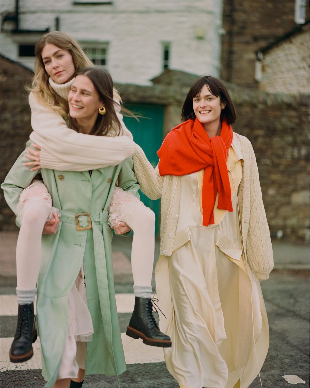 Three women in fashionable coats and dresses walking outdoors; one woman is giving another a piggyback ride on a city street.